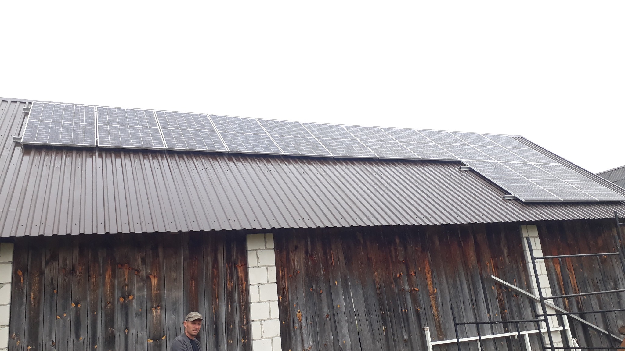 Panele słoneczne zamontowane na dachu starej stodoły z drewnianymi ścianami i blachodachówką, widoczny mężczyzna w czapce i drabina oparta o budynek.
