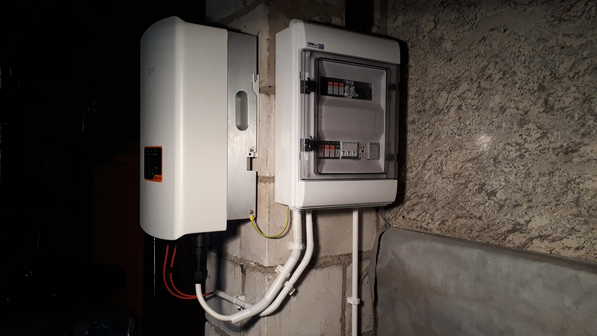 Biały inwerter fotowoltaiczny oraz skrzynka elektryczna z bezpiecznikami zamontowane na ścianie z cegły i betonu wewnątrz pomieszczenia.