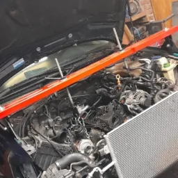 Silnik samochodowy z otwartą maską, podtrzymywany pomarańczową belką, widoczny chłodnica i inne elementy w trakcie naprawy w warsztacie.