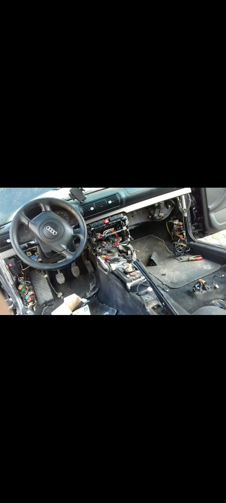 Wnętrze samochodu Audi A4 w trakcie demontażu deski rozdzielczej, widoczne liczne przewody i elementy instalacji elektrycznej, kierownica z logo Audi po lewej stronie.