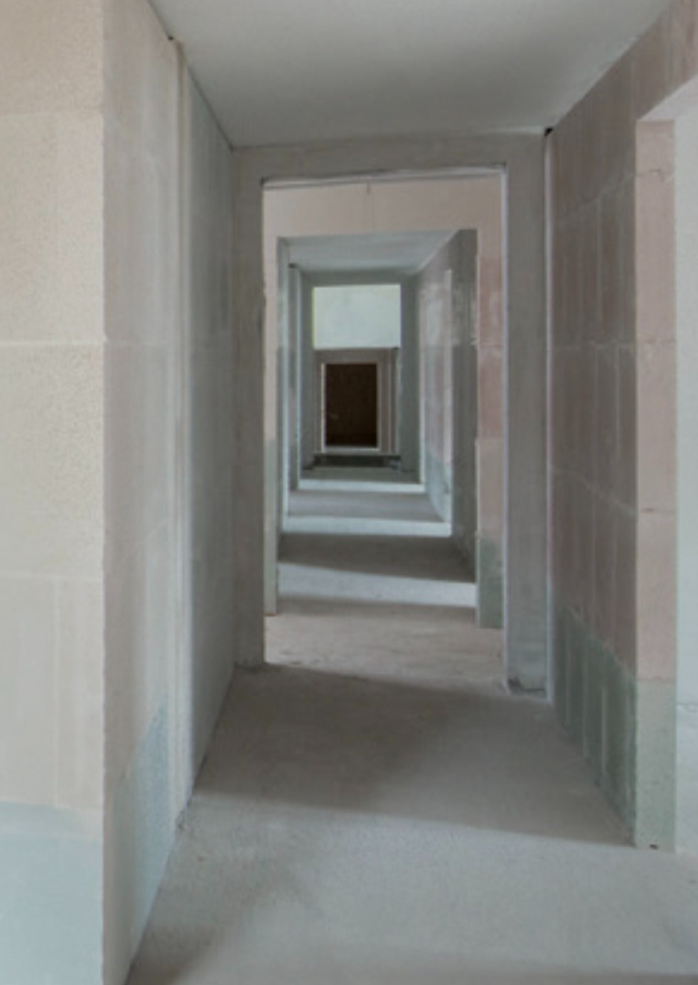 Długi korytarz w stanie surowym, z widocznymi surowymi ścianami z bloków i betonową podłogą, perspektywa zbiegająca się do ciemnych drzwi na końcu korytarza.