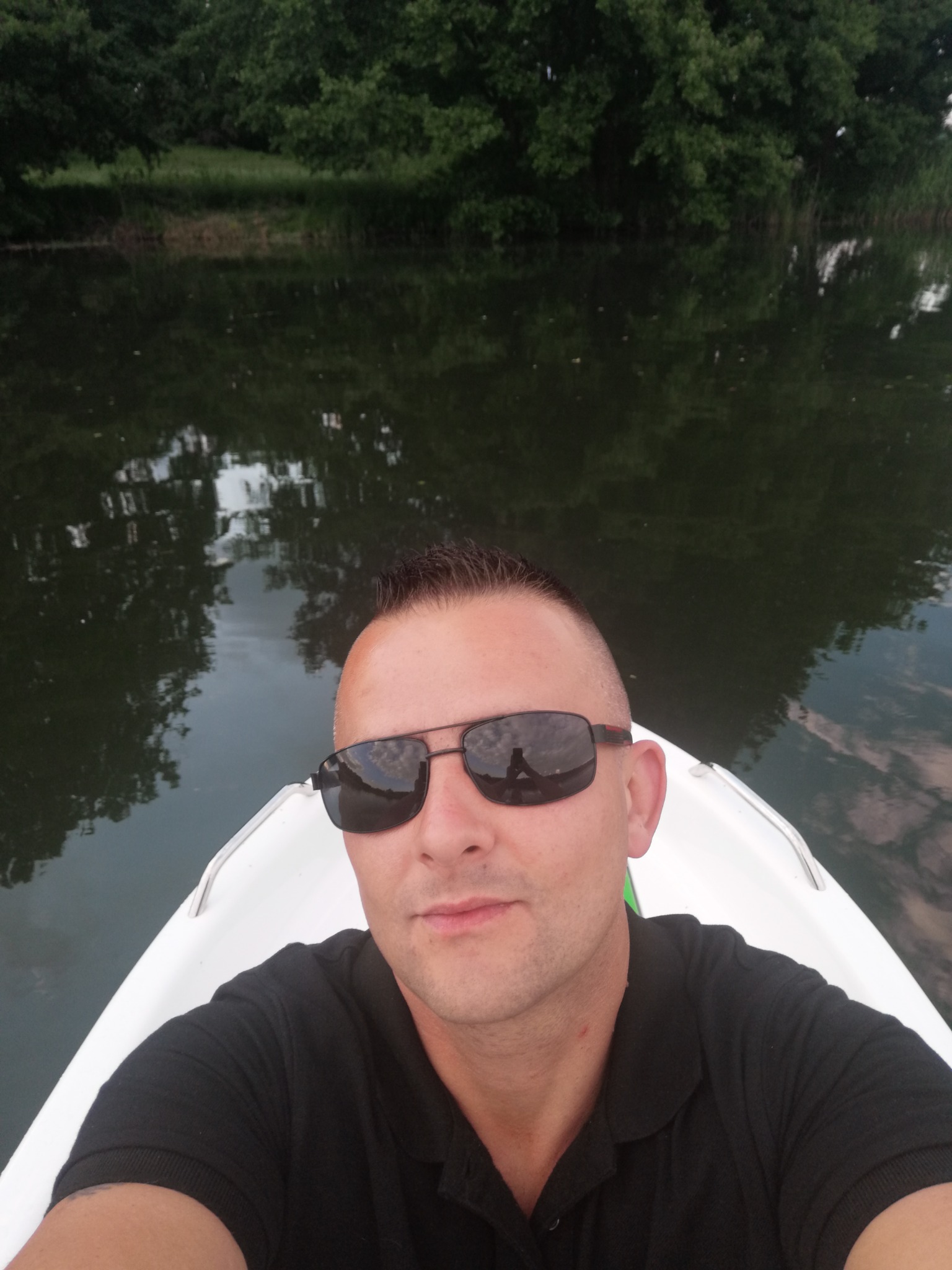 Mężczyzna w czarnej koszulce polo i okularach przeciwsłonecznych robi selfie na łódce na tle jeziora i zieleni.