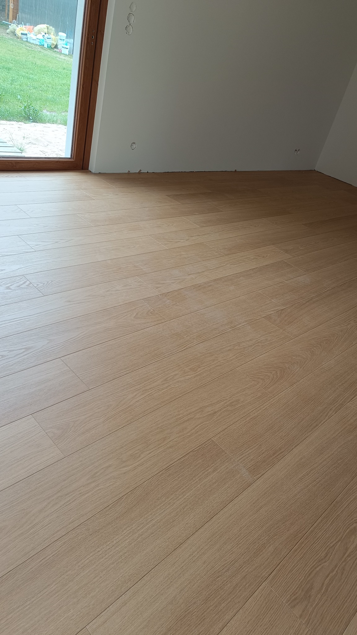 Jasne panele podłogowe w pokoju na poddaszu z oknem. Widoczne puszki elektryczne na ścianie, co sugeruje świeżo wykończone wnętrze.