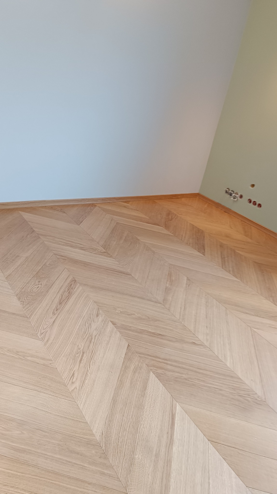 Jasny parkiet ułożony we wzór jodełki francuskiej w rogu pokoju, widoczne listwy przypodłogowe i gniazdka elektryczne na ścianie. Naturalne światło podkreśla strukturę drewna.