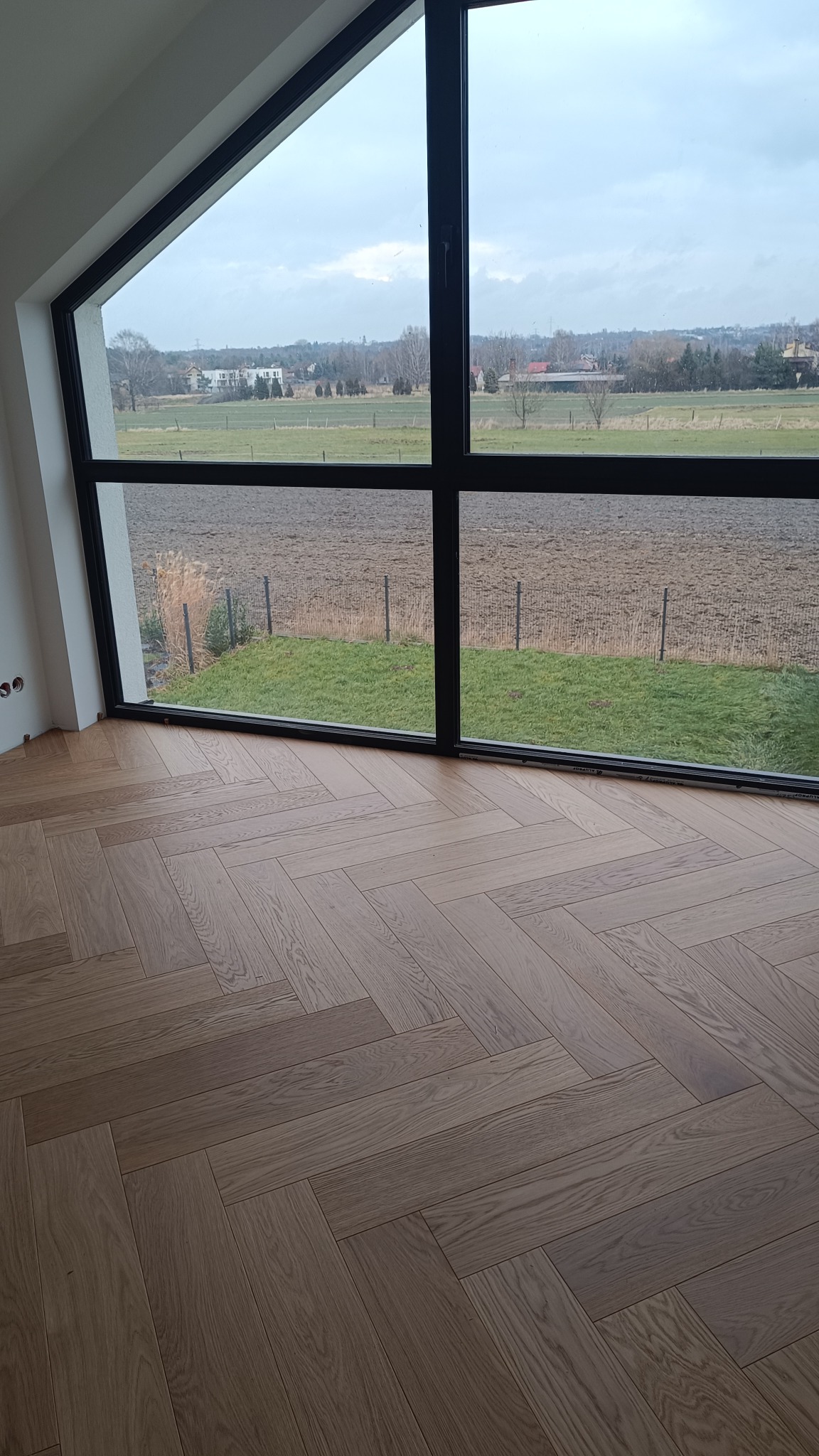 Jasny parkiet ułożony w jodełkę francuską, widoczny przez duże okno z czarnymi ramami i widokiem na pole. Częściowo widoczne gniazdka elektryczne na ścianie.