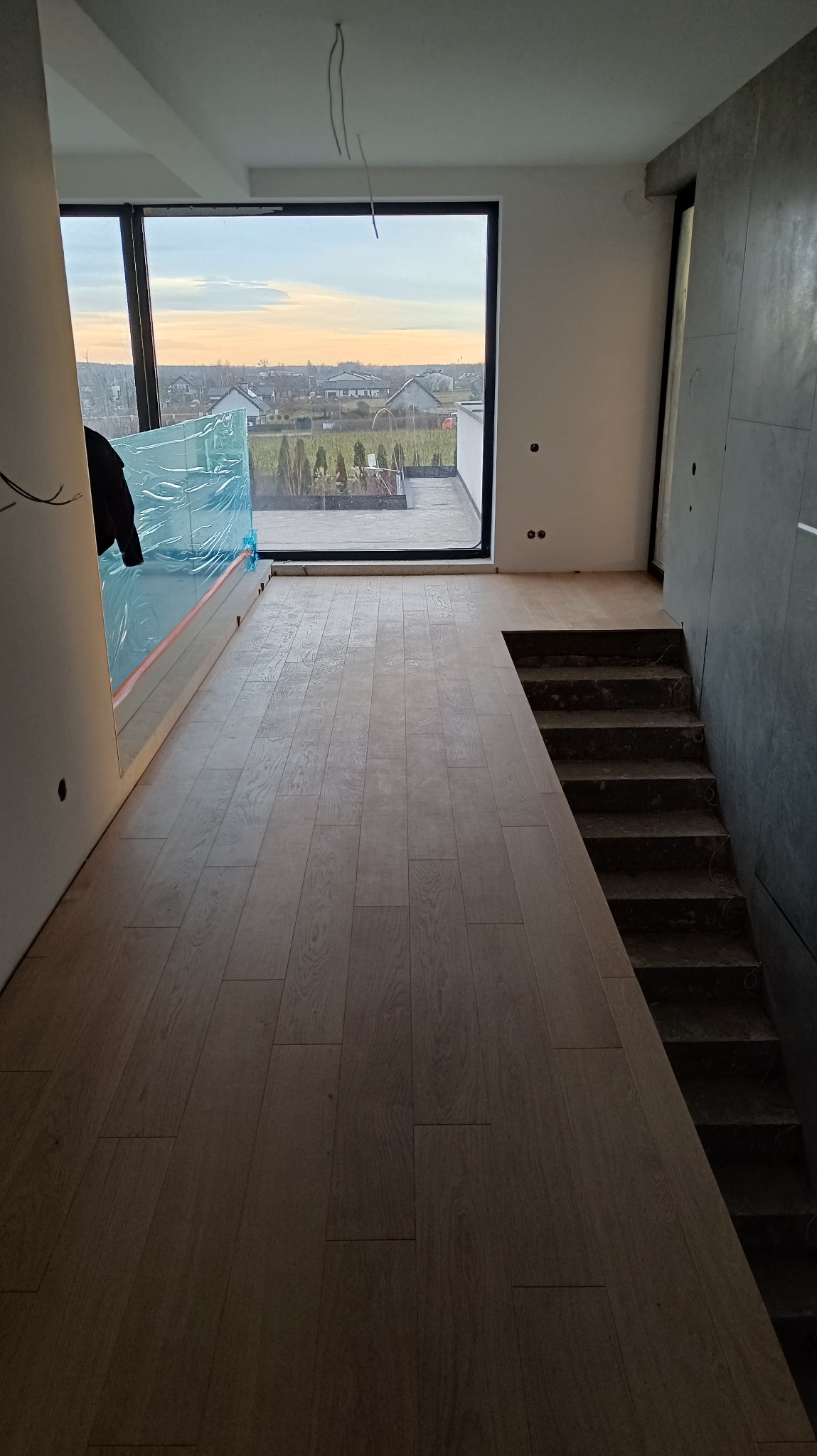 Jasne panele podłogowe ułożone w nowoczesnym wnętrzu z widokiem na zewnątrz przez duże okno. Schody w surowym stanie obok paneli. Część ściany zabezpieczona folią.