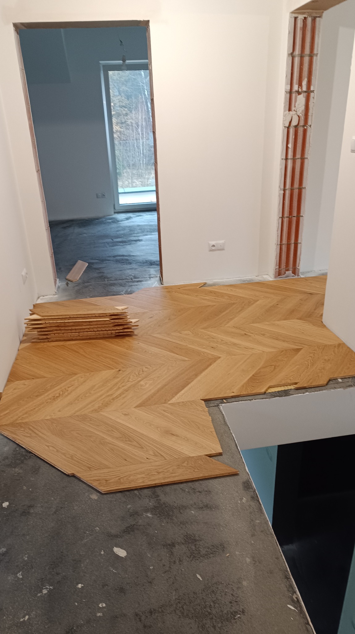 Układanie parkietu w jodełkę francuską. Częściowo ułożona podłoga z drewnianych desek, stos desek obok. Widoczne przygotowane podłoże i fragment ściany.