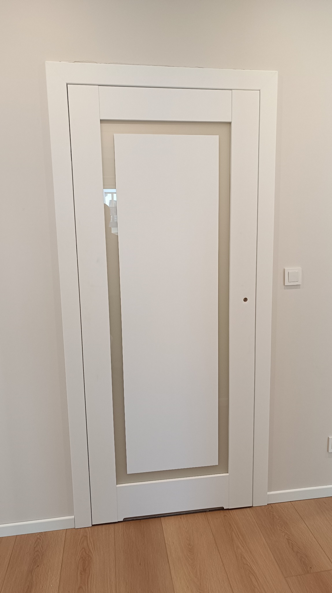 Białe, nowoczesne drzwi wewnętrzne z przeszkleniem, widoczne na tle jasnej ściany i podłogi z paneli. Minimalistyczny design, proste linie.