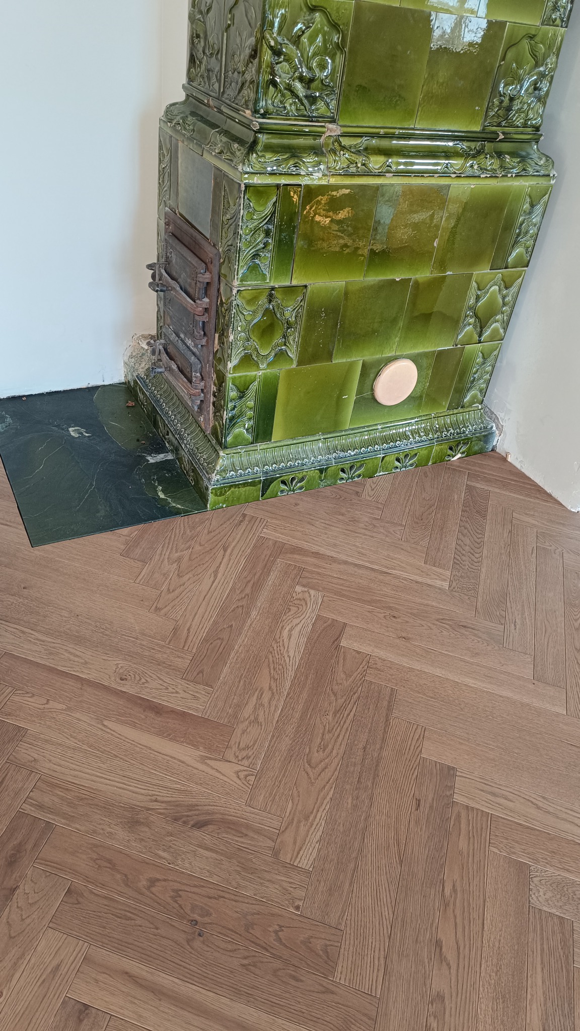 Ułożony parkiet w jodełkę w odcieniach brązu, sąsiadujący z ozdobnym, zielonym piecem kaflowym. Widok na fragment podłogi i piec, z uwzględnieniem detali wzoru parkietu.