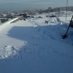 Śnieg na płaskim dachu z widocznymi śladami odśnieżania, w tle zabudowa miejska i błękitne niebo.