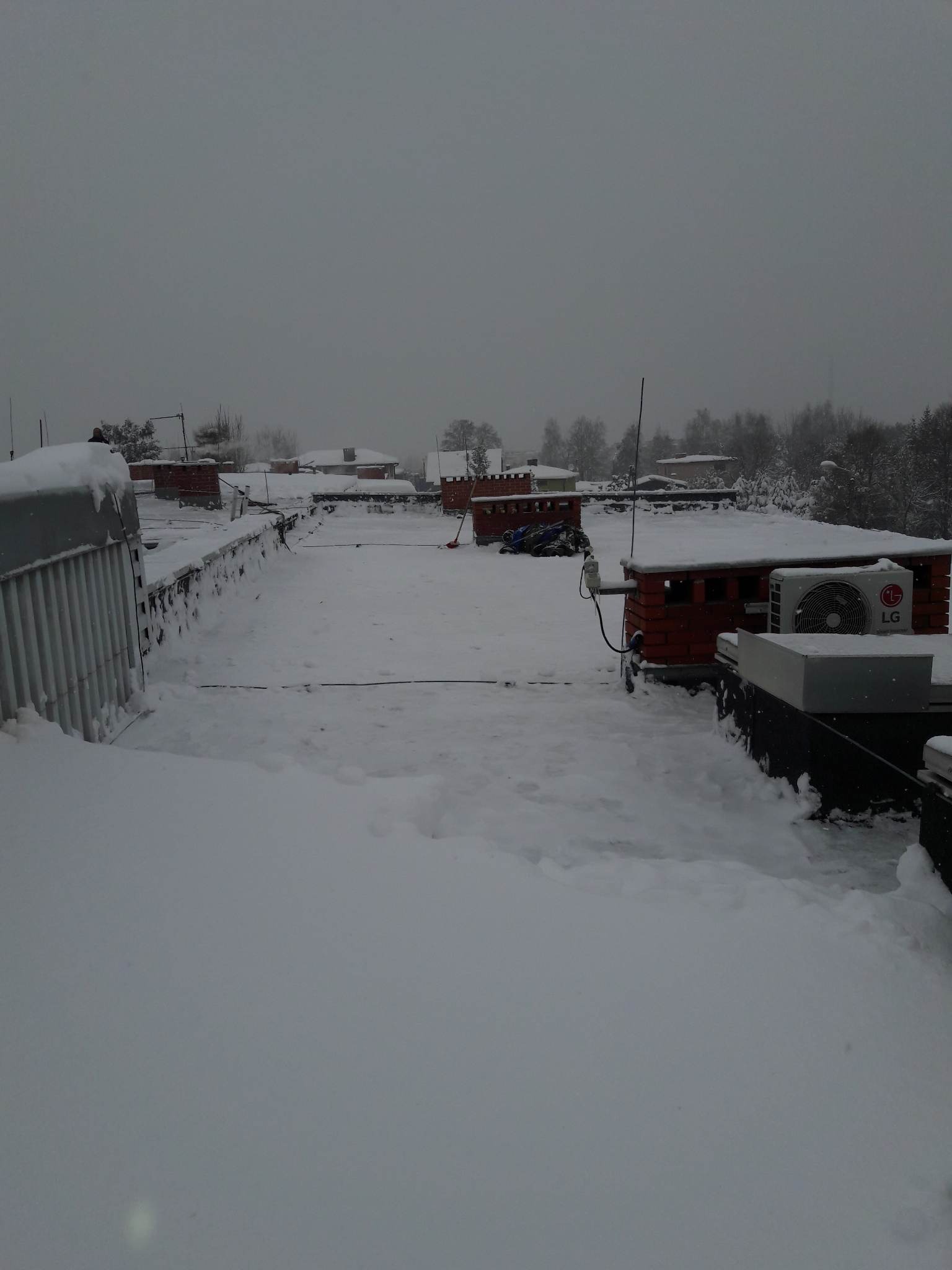 Zasypany śniegiem płaski dach budynku z widocznym agregatem klimatyzacji LG, anteną i czerwoną łopatą do śniegu w tle zabudowa mieszkalna.