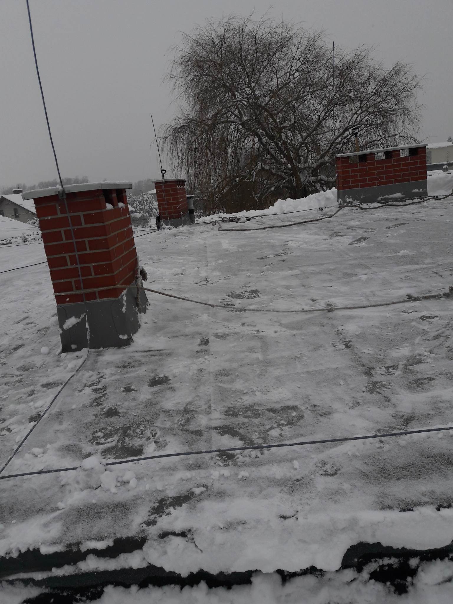 Śnieżny dach z ceglanymi kominami i przewodami odgromowymi, w tle drzewo bez liści i zabudowania w zimowej scenerii.