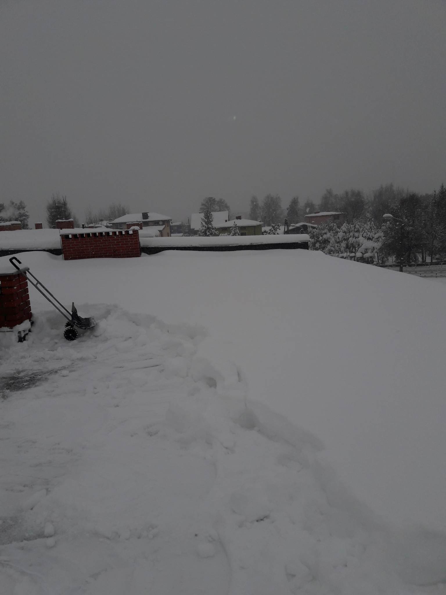 Zaśnieżony dach z odgarniętą częścią śniegu, widoczny komin i odśnieżarka ręczna na tle pochmurnego nieba.