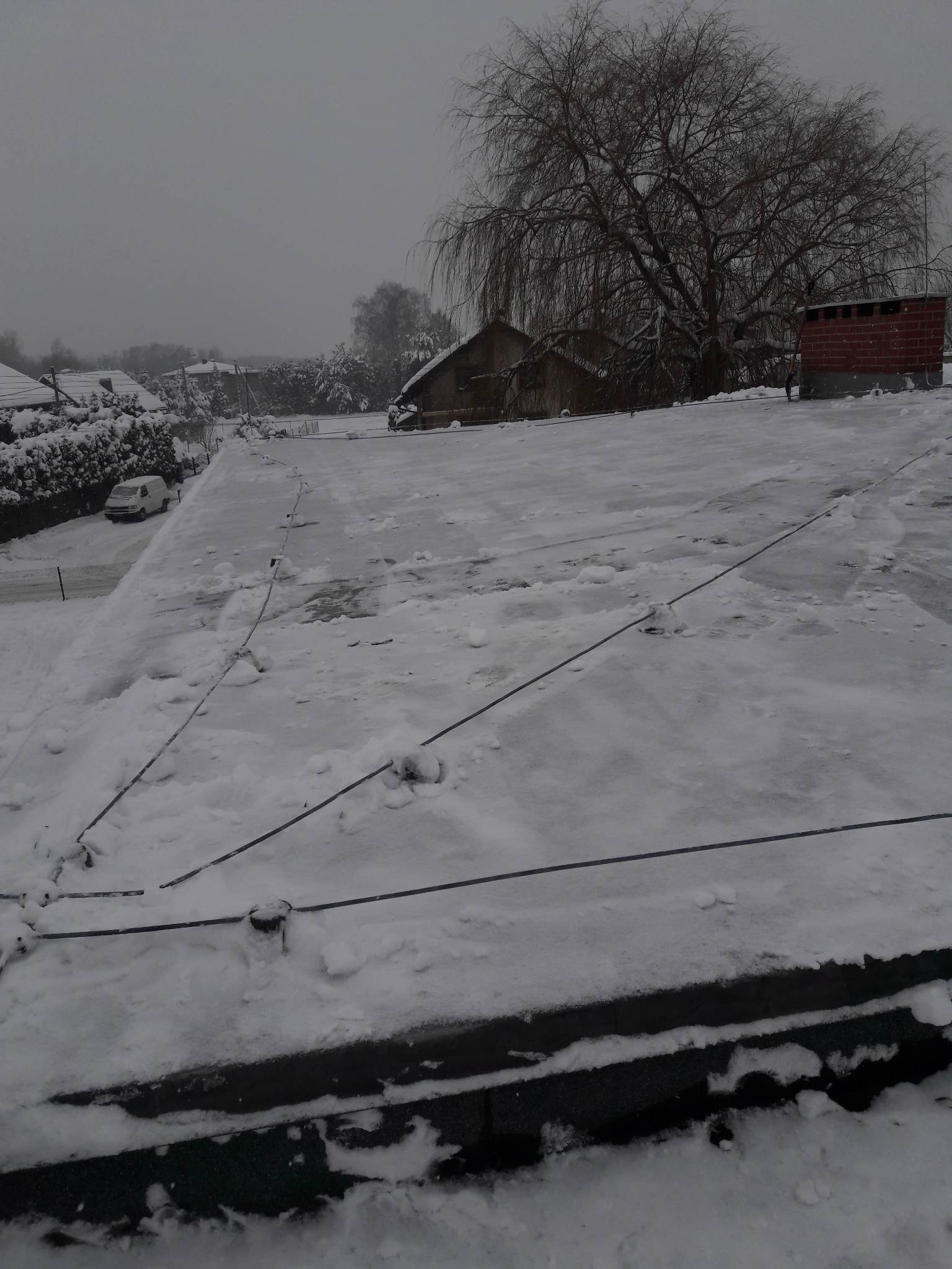 Śnieg na płaskim dachu z widocznymi elementami instalacji odgromowej, w tle budynki i drzewo bez liści w zimowej scenerii.