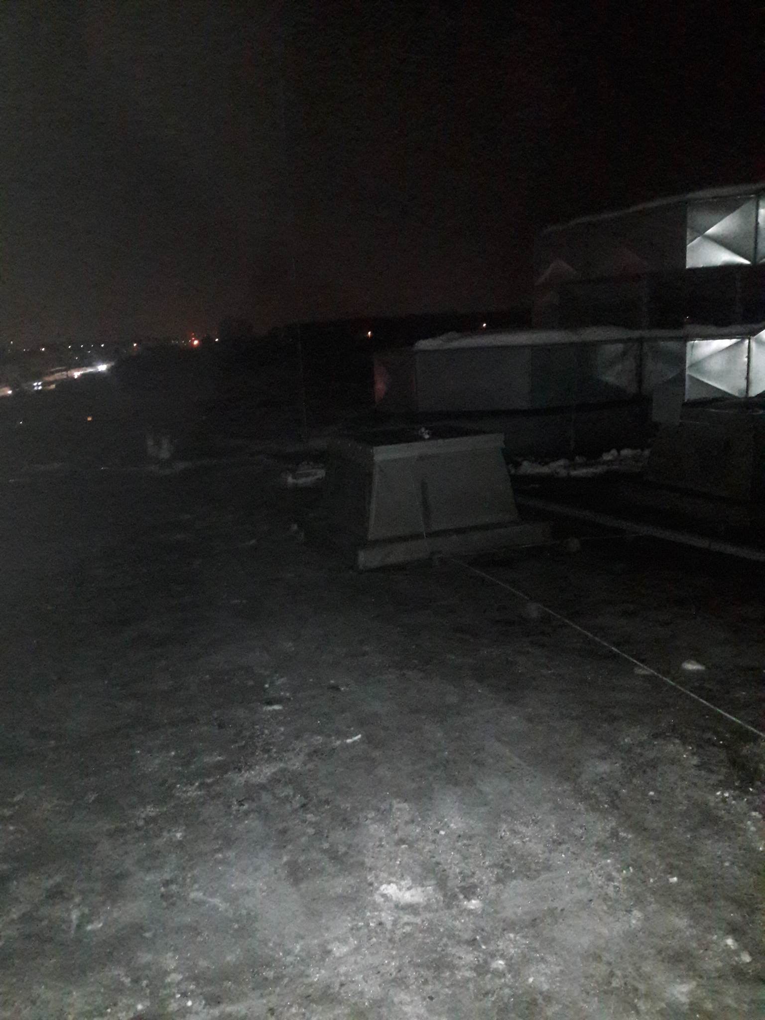 Nocny widok na płaski dach pokryty resztkami śniegu, na którym widoczne są elementy instalacji wentylacyjnej i w oddali światła miasta.