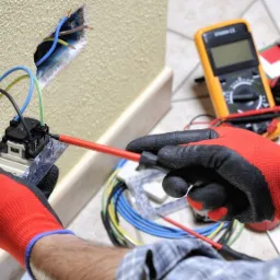 Elektryk w czerwonych rękawicach montuje gniazdko elektryczne w ścianie, używając śrubokręta; widoczne przewody, multimetr, notatnik i taśma miernicza.