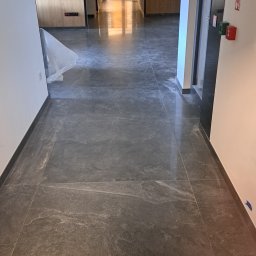 Prace wykończeniowe bracia - Długi korytarz w budynku w trakcie wykończenia, z szarą, marmurkową posadzką z płytek, zabezpieczoną folią fragmentem ściany, widoczne drzwi i oświetlenie sufitowe.