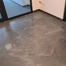 Nowoczesna, szara podłoga z płytek imitujących kamień, ułożona w nowym pomieszczeniu z białymi ścianami i czarnymi ramami okiennymi, widoczne gniazdka elektryczne.