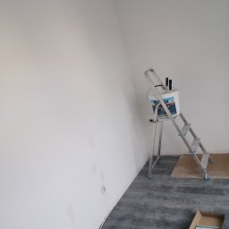 Prace wykończeniowe bracia - Świeżo pomalowana biała ściana z widocznymi smugami po malowaniu, aluminiowa drabina z wiadrem farby i pędzlami, oraz fragment podłogi z panelami w trakcie układania.