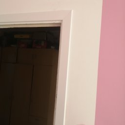 Prace wykończeniowe bracia - Fragment ościeżnicy pomalowanej na biało, przylegającej do różowej ściany. W tle widoczne wnętrze szafy wypełnionej pudełkami.