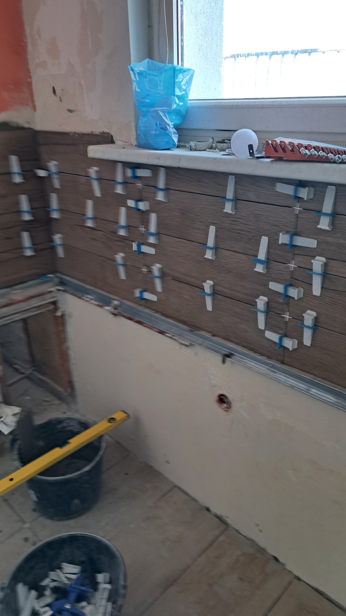 Układanie płytek imitujących drewno na ścianie, widoczne klipsy poziomujące, poziomica oparta o wiadro z zaprawą, okno w tle.