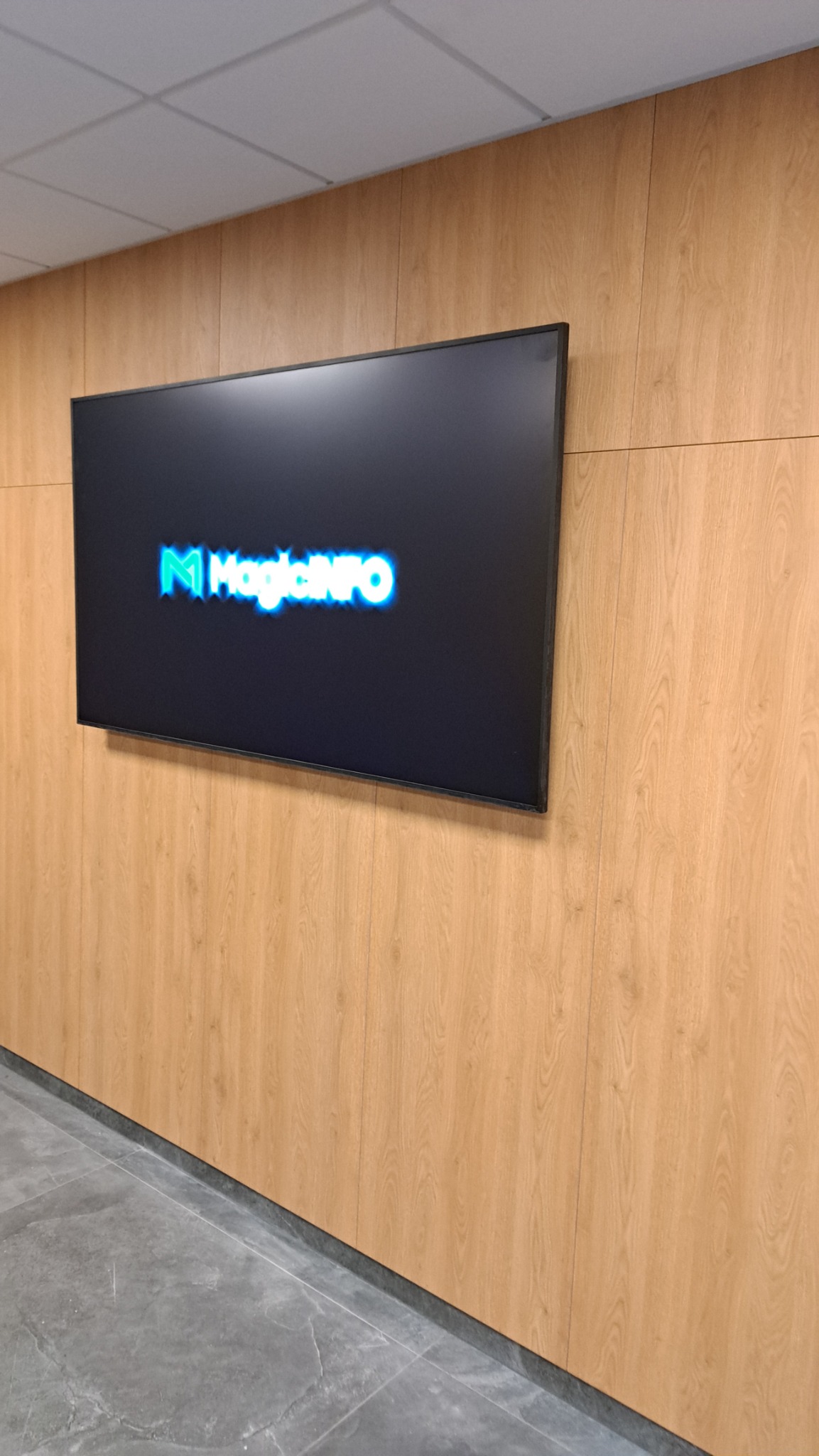 Telewizor z logiem MagicINFO zamontowany na ścianie wyłożonej panelami imitującymi drewno.