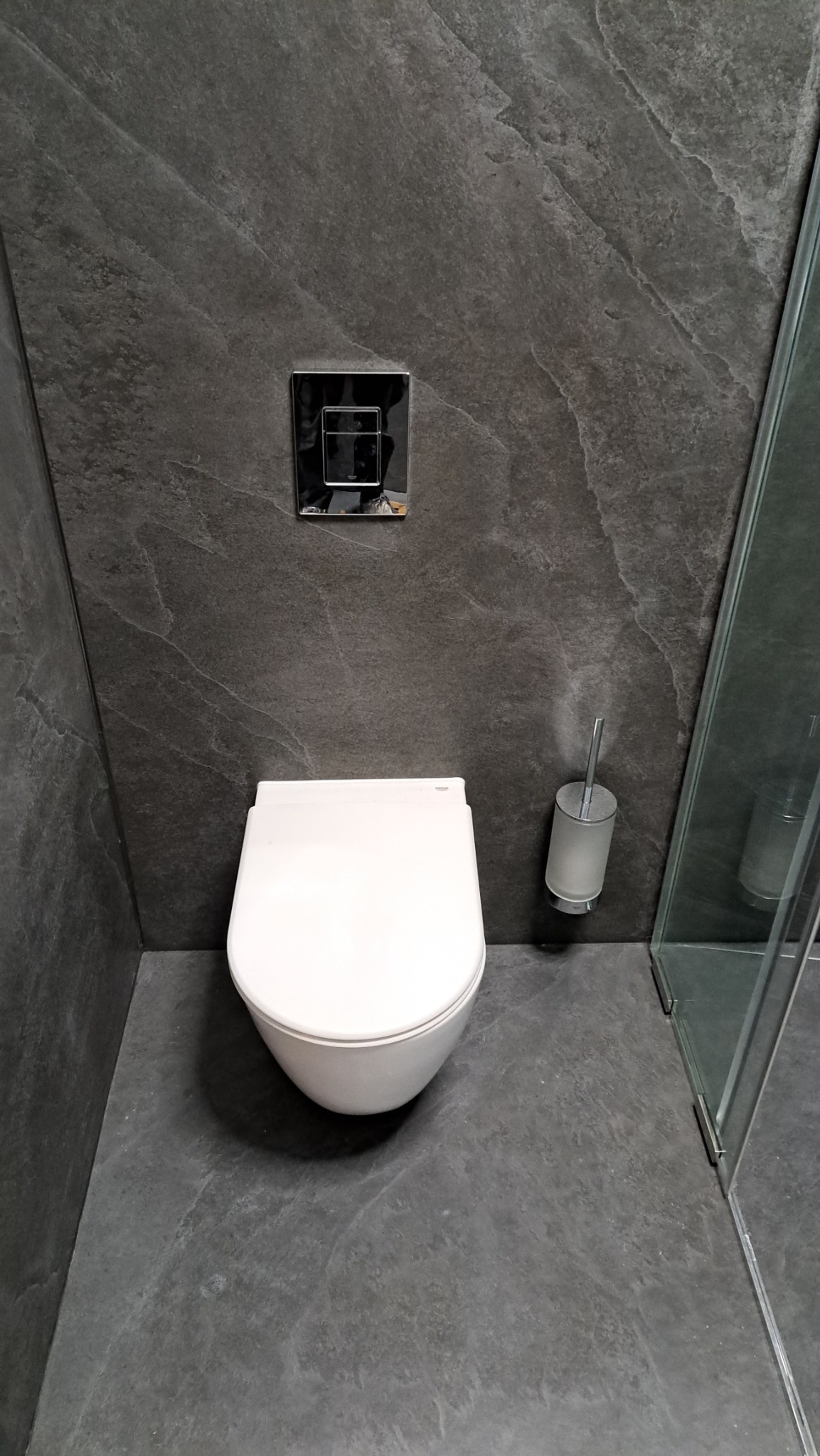 Nowoczesna toaleta podwieszana z chromowanym przyciskiem spłukującym na ścianie z szarej imitacji kamienia, obok szczotka do WC i kabina prysznicowa