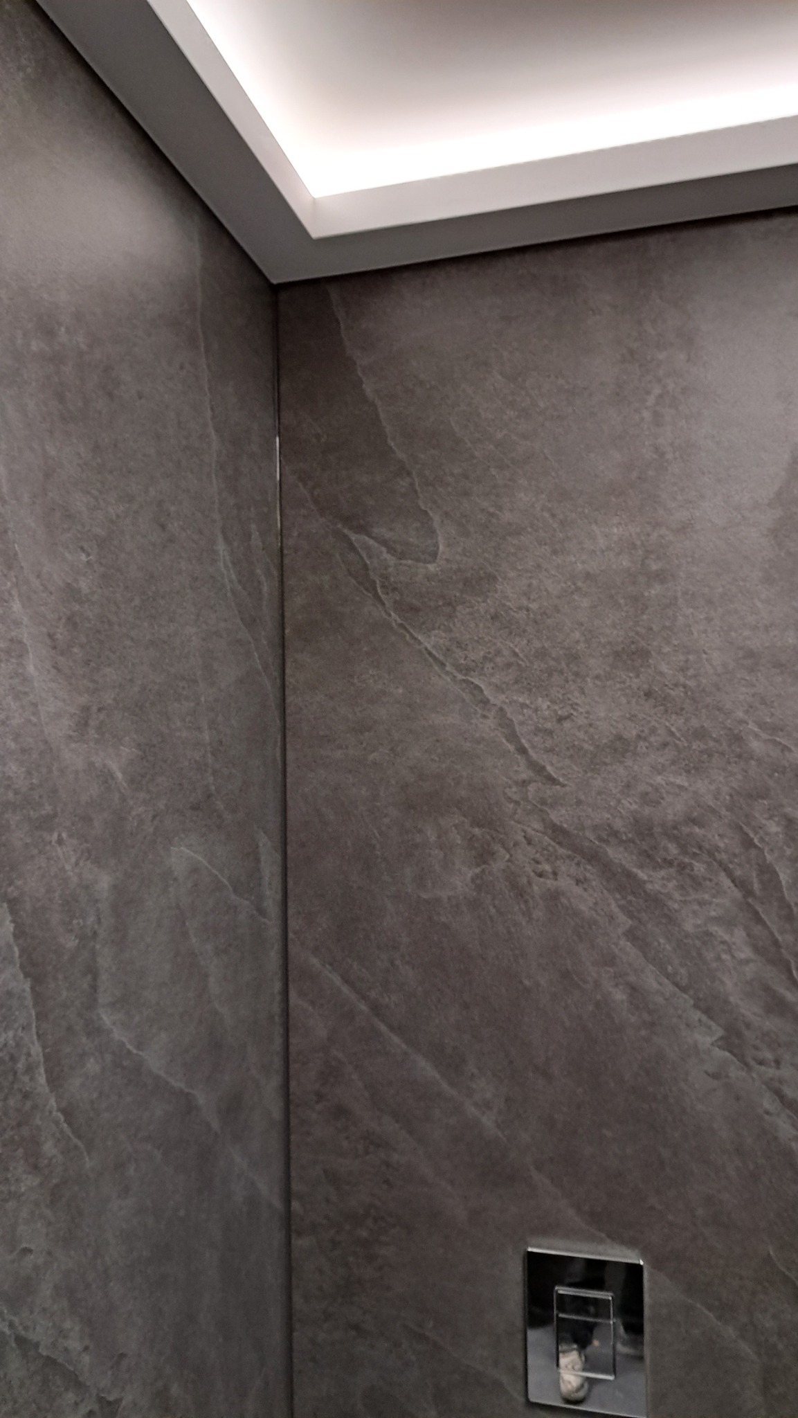 Szare płyty imitujące kamień na ścianie w łazience, widoczny fragment sufitu z podświetleniem LED i bateria podtynkowa.