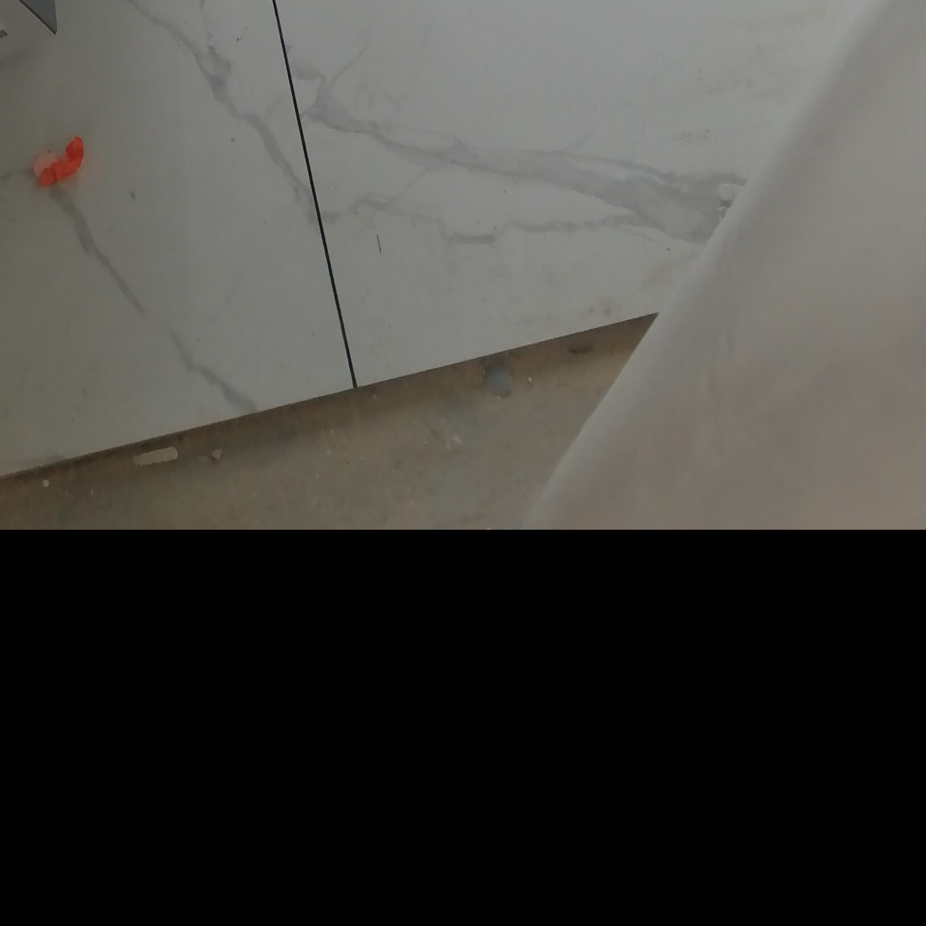 Fragment ułożonej podłogi z białych płytek imitujących marmur, widoczny pomarańczowy krzyżyk dystansowy.