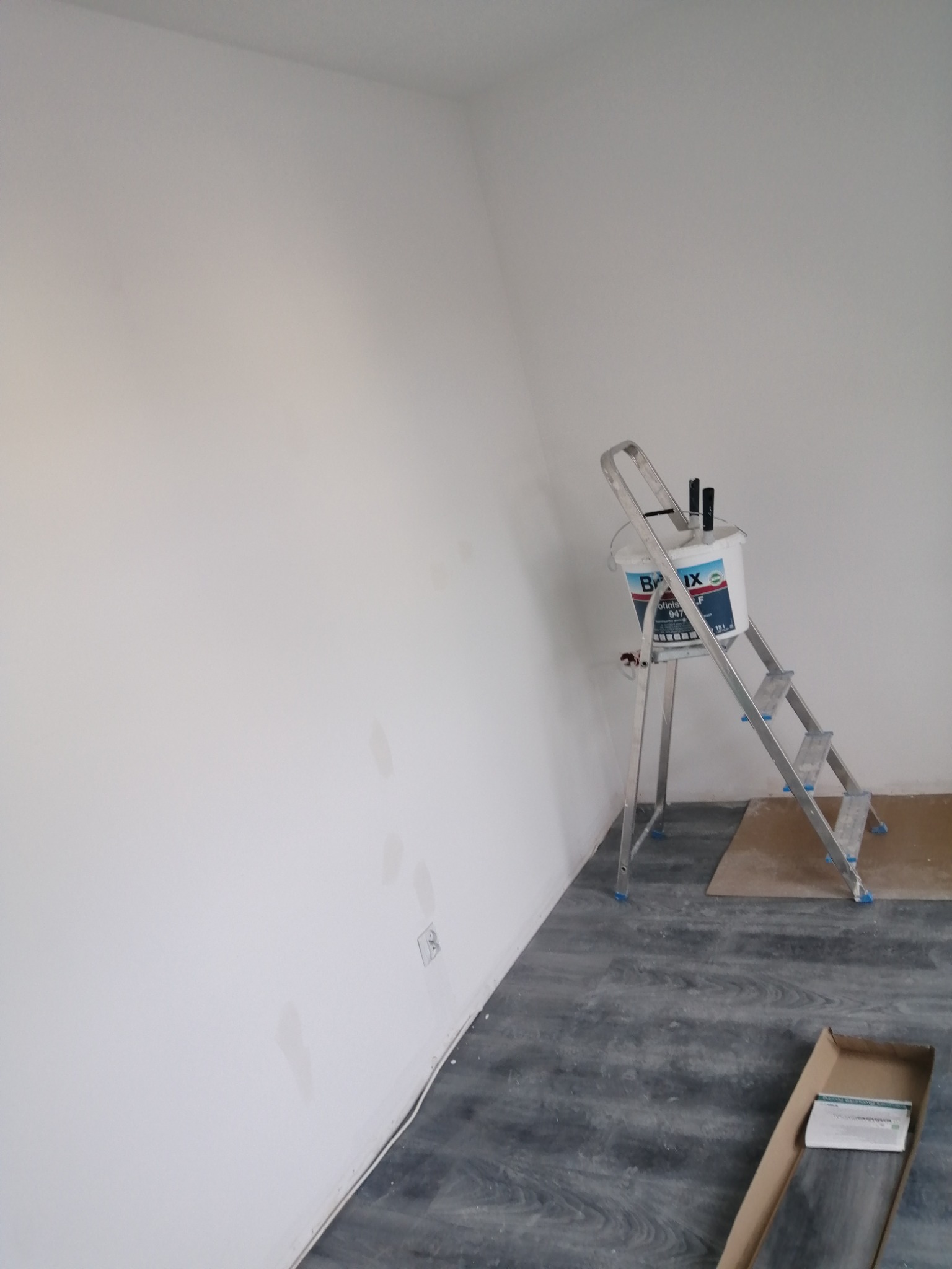 Świeżo pomalowana biała ściana z widocznymi smugami po malowaniu, aluminiowa drabina z wiadrem farby i pędzlami, oraz fragment podłogi z panelami w trakcie układania.