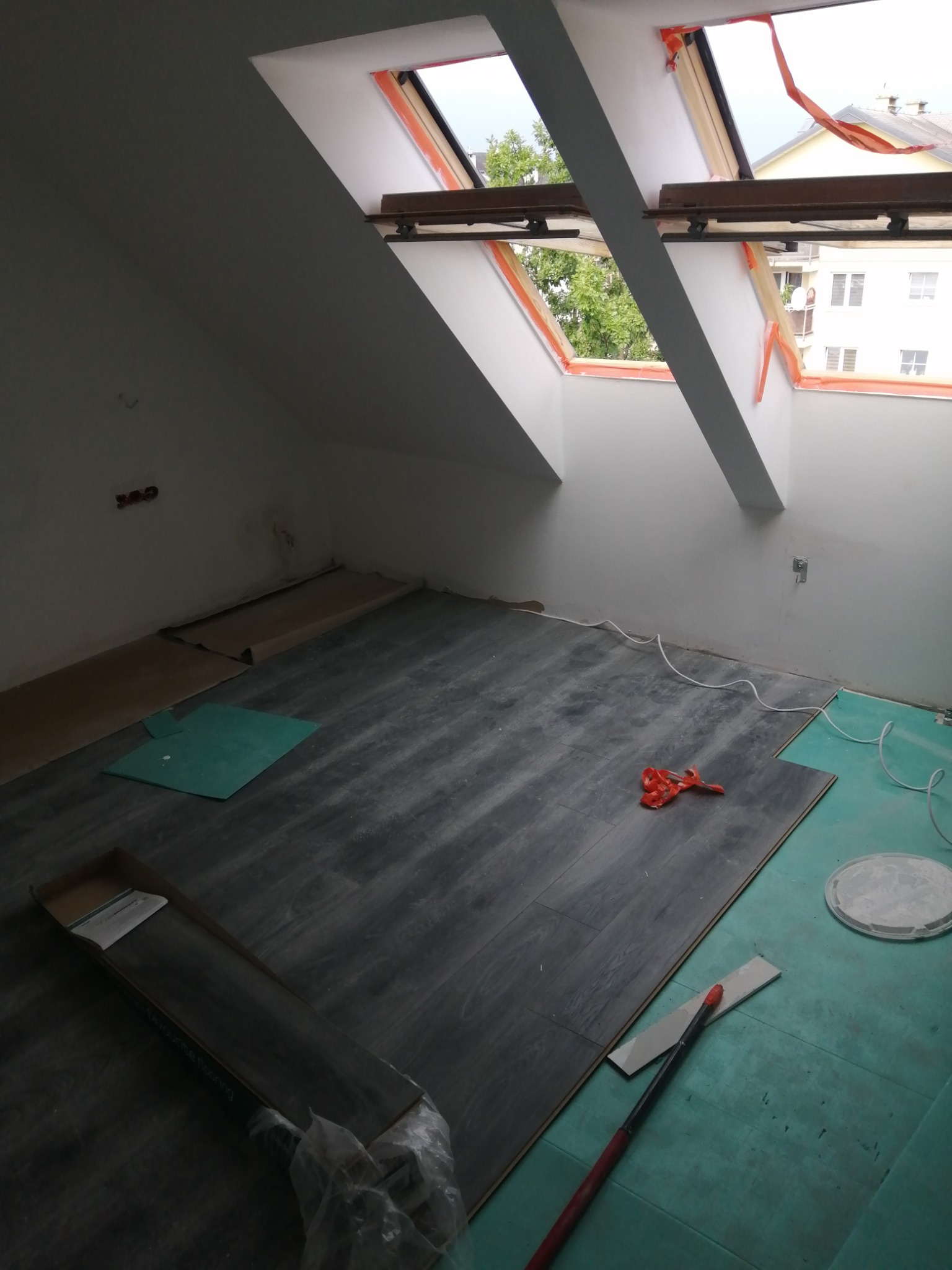 Montaż paneli podłogowych w pokoju na poddaszu z oknami dachowymi, widoczne narzędzia, podkład i częściowo ułożona podłoga w odcieniach szarości.