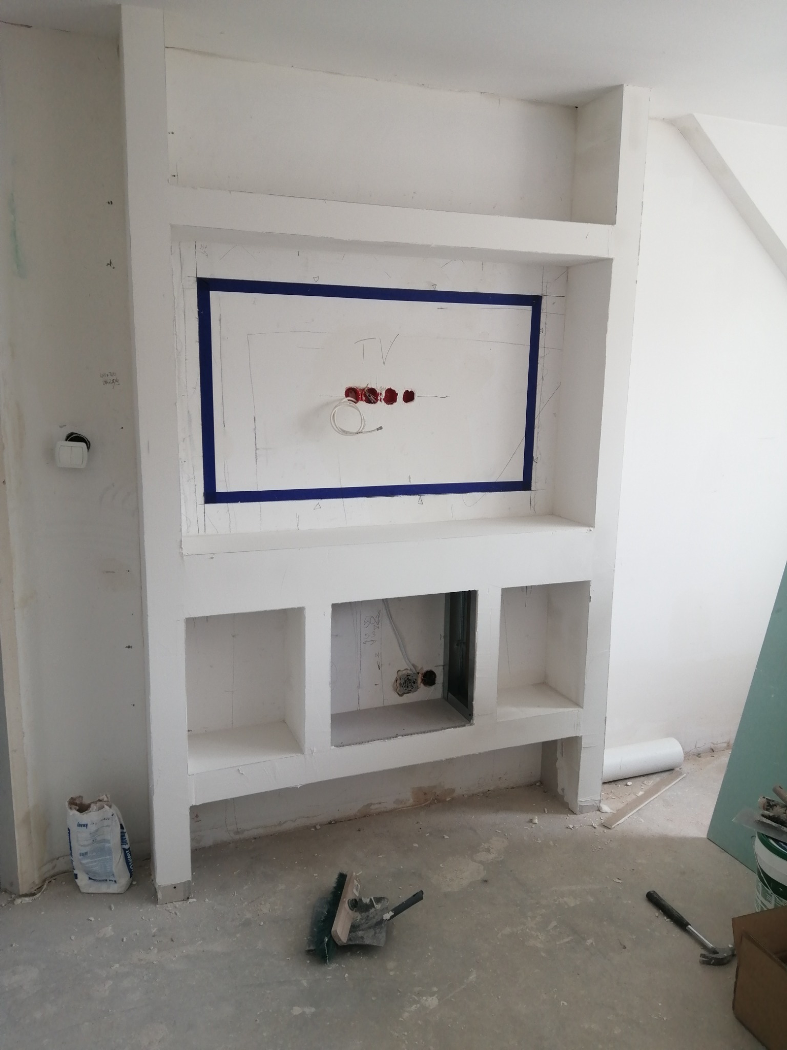 Niewykończona zabudowa z płyt gipsowo-kartonowych, z wnęką na telewizor obrysowaną niebieską taśmą, widoczne kable elektryczne, szpachla na podłodze i narzędzia.