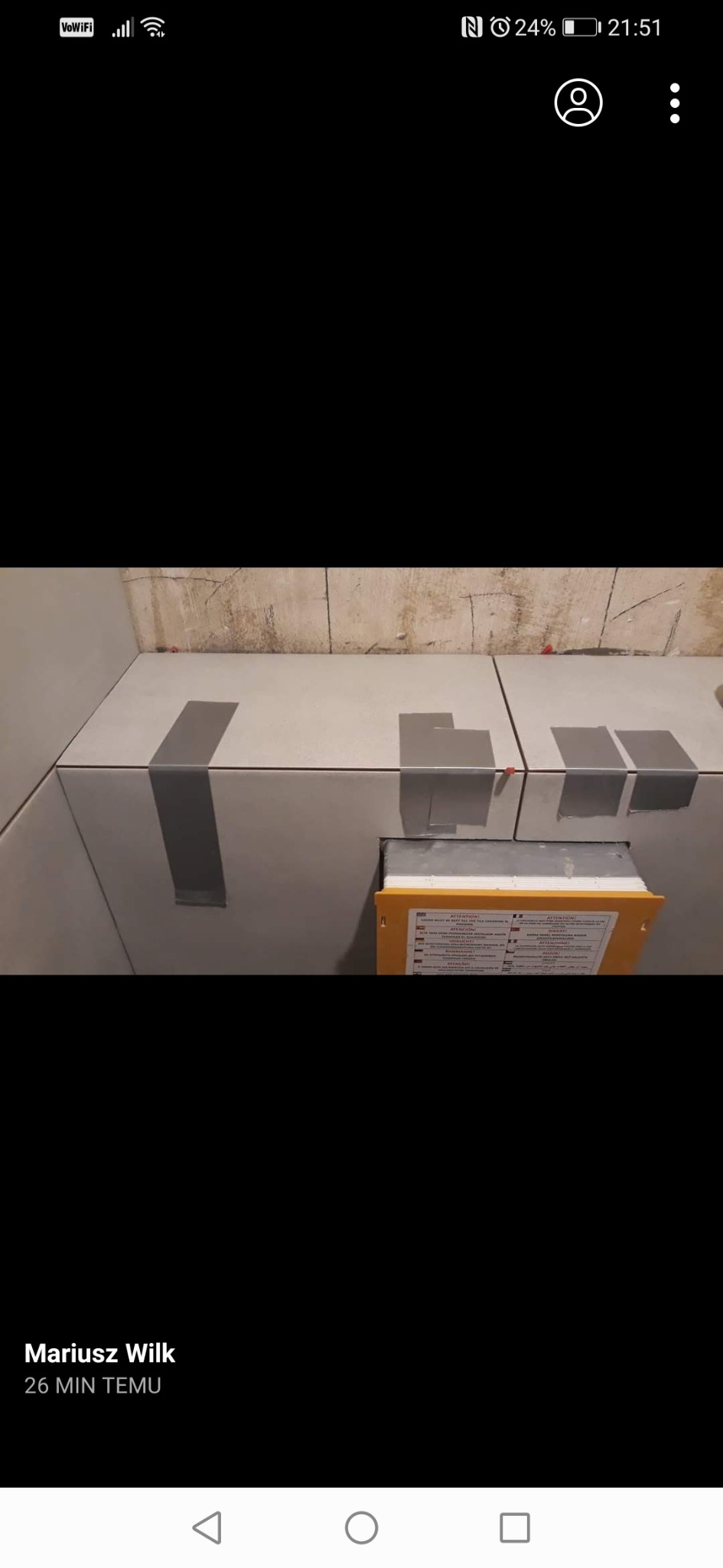 Szare płytki ścienne w trakcie montażu, zabezpieczone taśmą malarską, z widocznym dystanserem i instrukcją w żółtej obudowie.