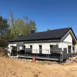 Dom jednorodzinny w trakcie budowy, z widocznym ociepleniem ścian zewnętrznych i nowym, ciemnym dachem z kominem, otoczony rusztowaniami i ziemią.