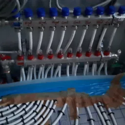 Rozdzielacz ogrzewania podłogowego z niebieskimi i czerwonymi zaworami, widoczne białe rurki i podłączenia do instalacji w podłodze, zabezpieczone niebieską folią.
