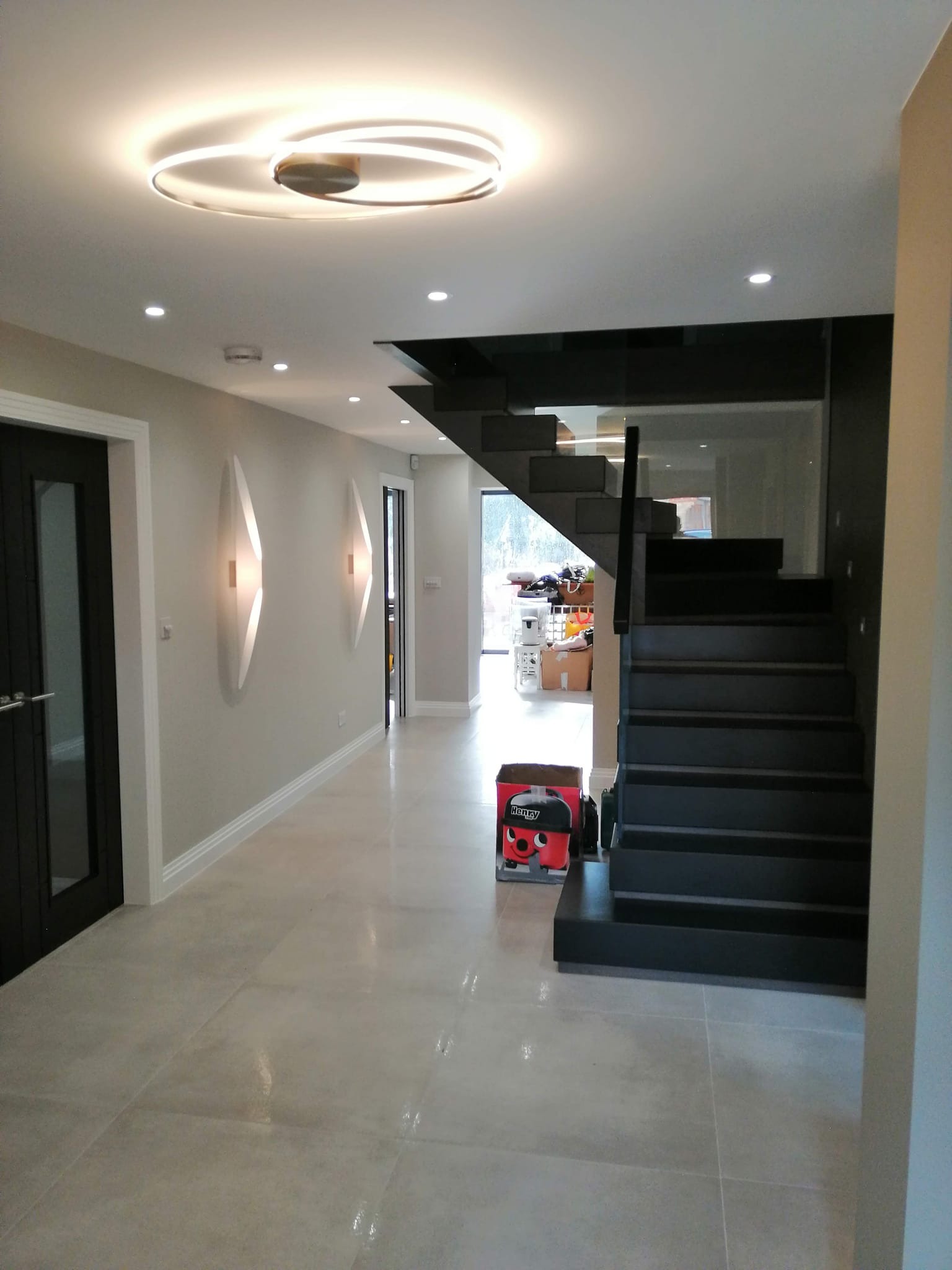 Nowoczesny hol z czarnymi schodami, szarą podłogą z płytek, minimalistycznym oświetleniem i dekoracyjnymi lampami ściennymi w kształcie łez.