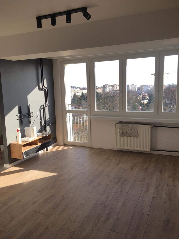 Jasne, przestronne wnętrze po remoncie z panelami podłogowymi imitującymi drewno, szarą ścianą z czarnymi rurami instalacyjnymi, białymi oknami i grzejnikami oraz widokiem na miasto za oknem.