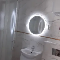 Nowoczesna łazienka z kabiną prysznicową, umywalką z szafką i okrągłym lustrem z podświetleniem LED na ścianie z marmurowym wzorem.