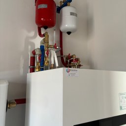 WeberKlima - Zainstalowana pompa ciepła Panasonic Aquarea Smart Cloud z czerwonym i białym zbiornikiem wyrównawczym, widoczne mosiężne rury i zawory, naklejka firmy Weberklima.