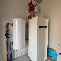 WeberKlima - Biała pompa ciepła Panasonic w kotłowni, z widocznymi rurami w kolorach czerwonym i niebieskim oraz zbiornikiem buforowym Galmet. Na ścianie zamontowane naczynia przeponowe w kolorach czerwonym...