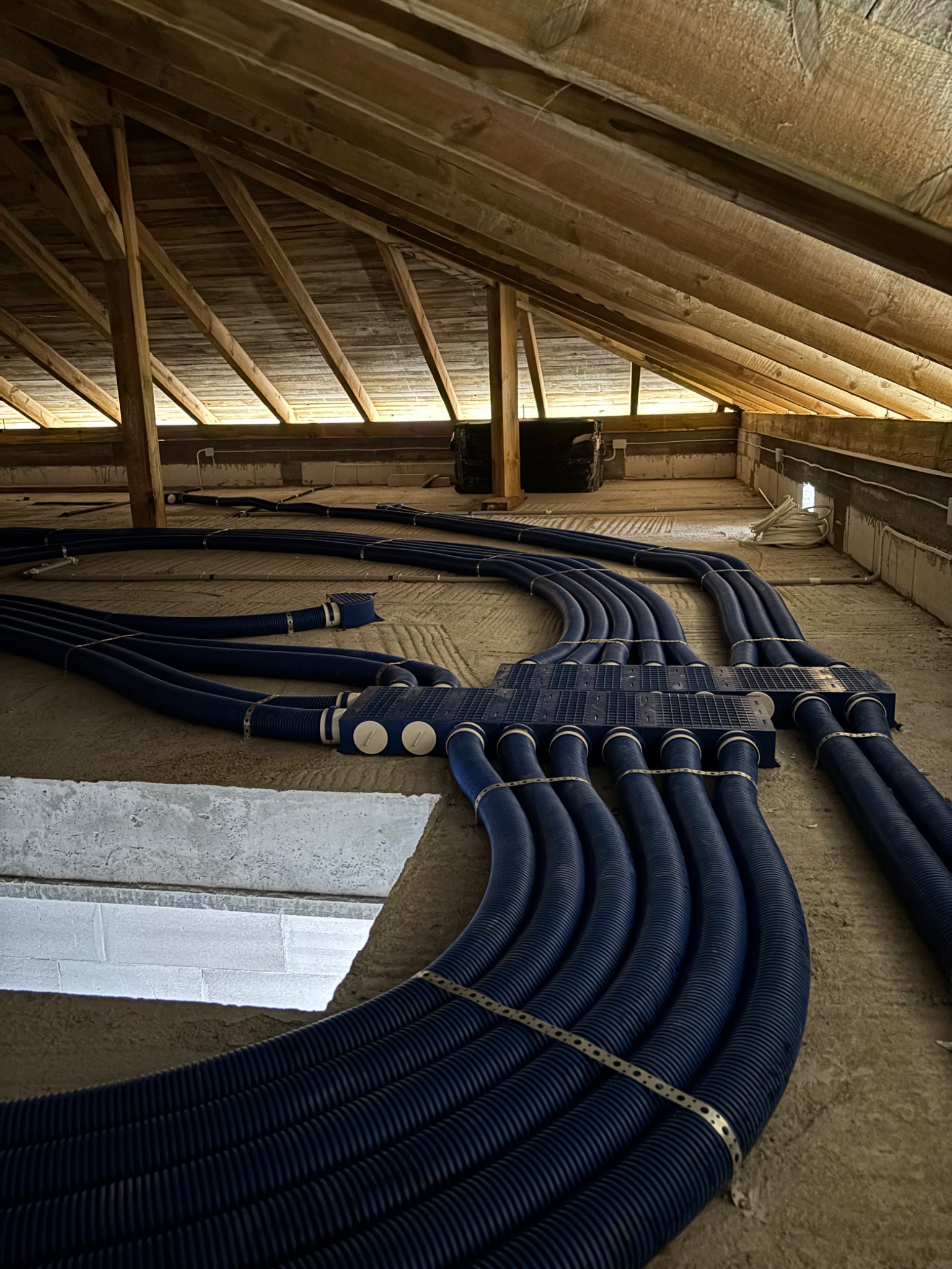 Niebieskie rury wentylacyjne, spięte metalowymi obejmami, rozchodzą się promieniście na poddaszu z drewnianą konstrukcją dachu.