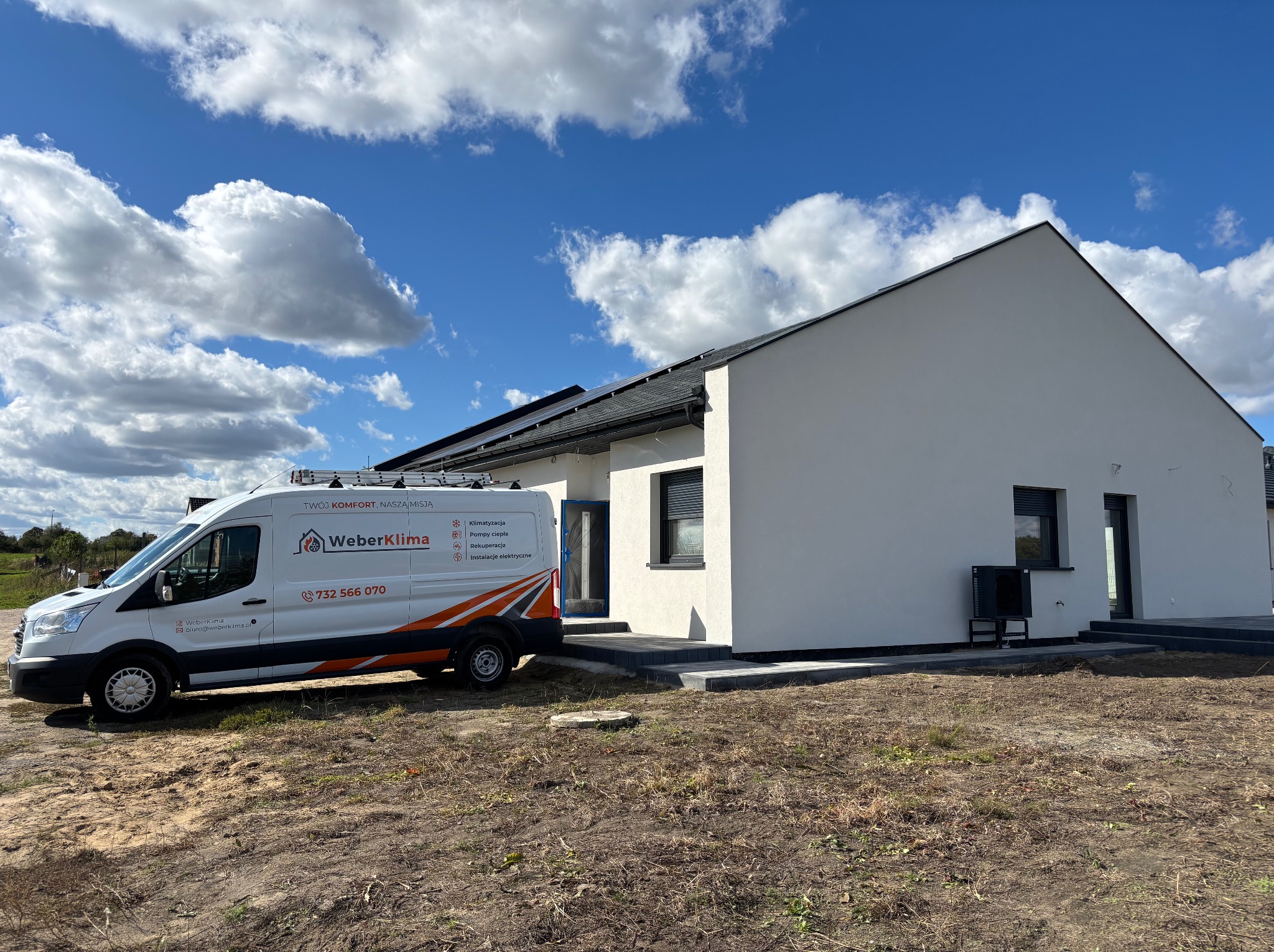 Biały budynek jednorodzinny z panelami słonecznymi na dachu, na tle błękitnego nieba z obłokami, z zaparkowanym obok samochodem dostawczym firmy WeberKlima z widocznym numerem telefonu i informacją...