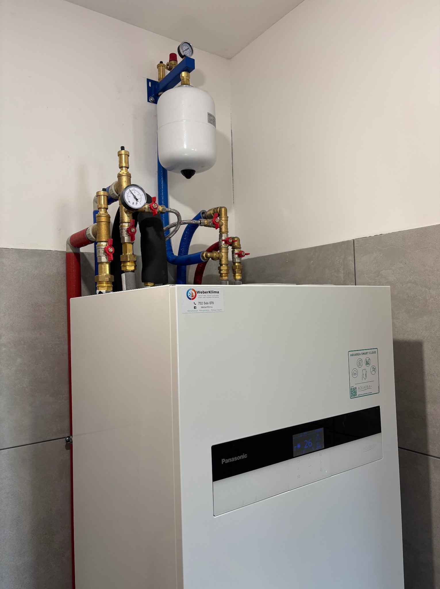 Biała pompa ciepła Panasonic z widocznymi rurami miedzianymi, zaworami i manometrami, zainstalowana w rogu pomieszczenia z szarymi płytkami. Na urządzeniu widoczna naklejka firmy Weberklima.