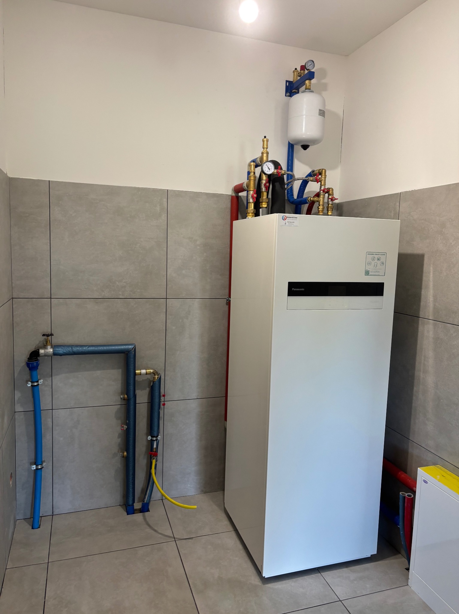 Biała pompa ciepła Panasonic zamontowana w pomieszczeniu z szarymi płytkami, widoczne przyłącza hydrauliczne z rurami w kolorach niebieskim i czerwonym oraz zbiornik wyrównawczy na górze,...