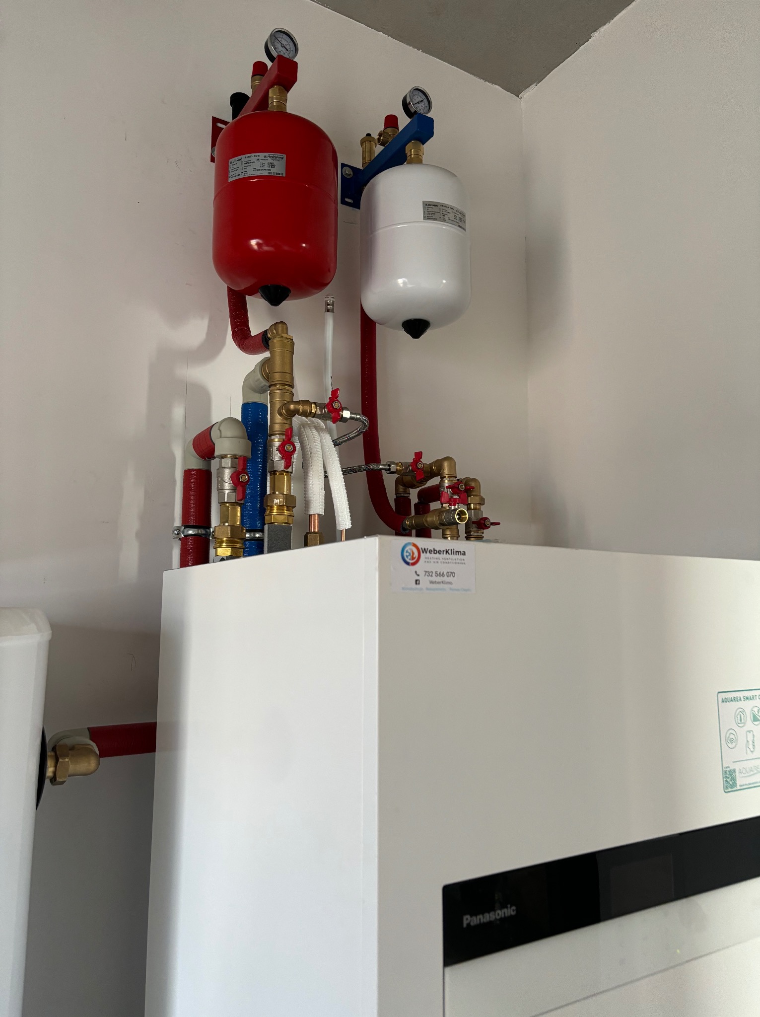 Zainstalowana pompa ciepła Panasonic Aquarea Smart Cloud z czerwonym i białym zbiornikiem wyrównawczym, widoczne mosiężne rury i zawory, naklejka firmy Weberklima.
