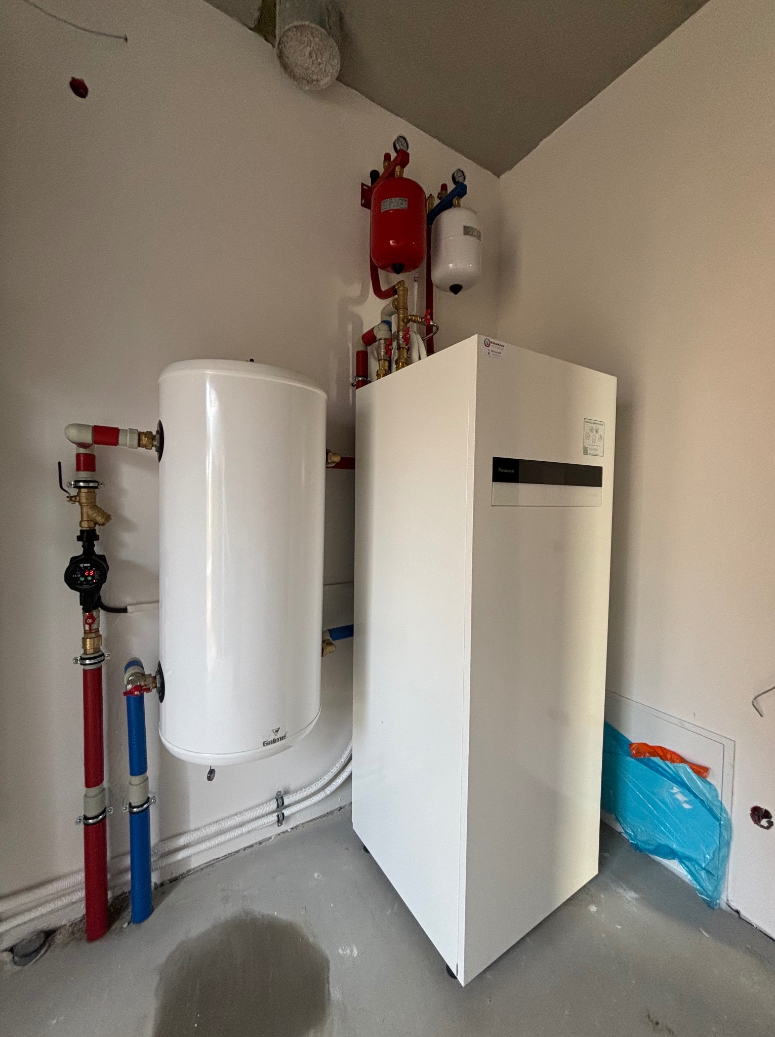 Biała pompa ciepła Panasonic w kotłowni, z widocznymi rurami w kolorach czerwonym i niebieskim oraz zbiornikiem buforowym Galmet. Na ścianie zamontowane naczynia przeponowe w kolorach czerwonym...