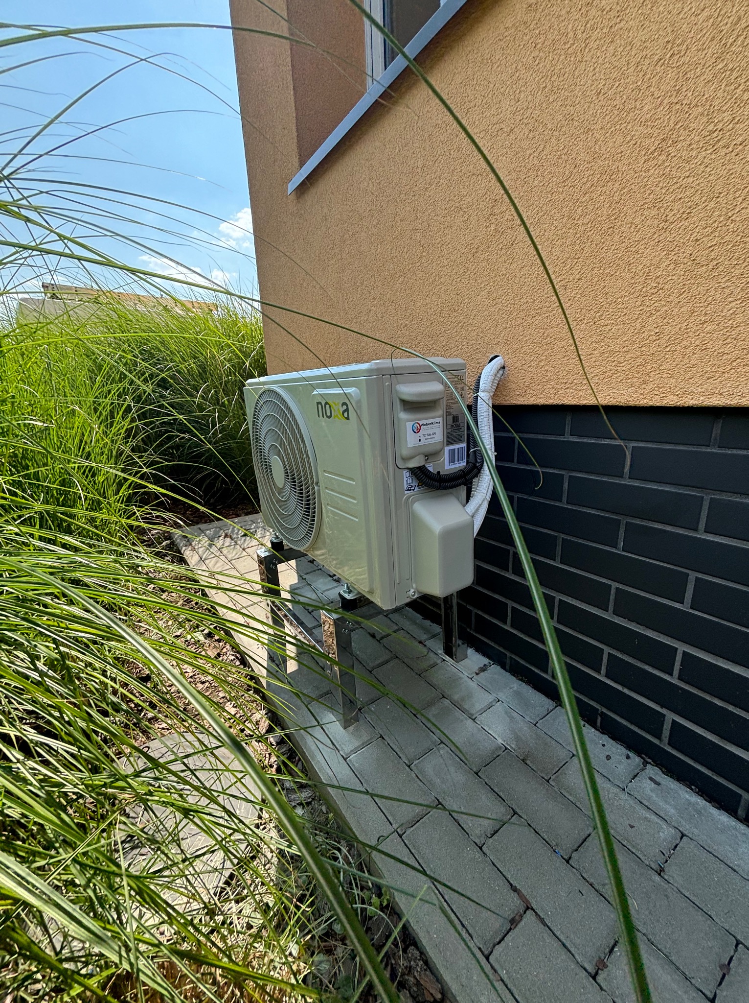 Zewnętrzna jednostka klimatyzacji marki Nooa zamontowana na metalowych wspornikach przy ścianie budynku z elewacją w kolorze piaskowym i cokole z ciemnej cegły, otoczona ozdobną trawą.