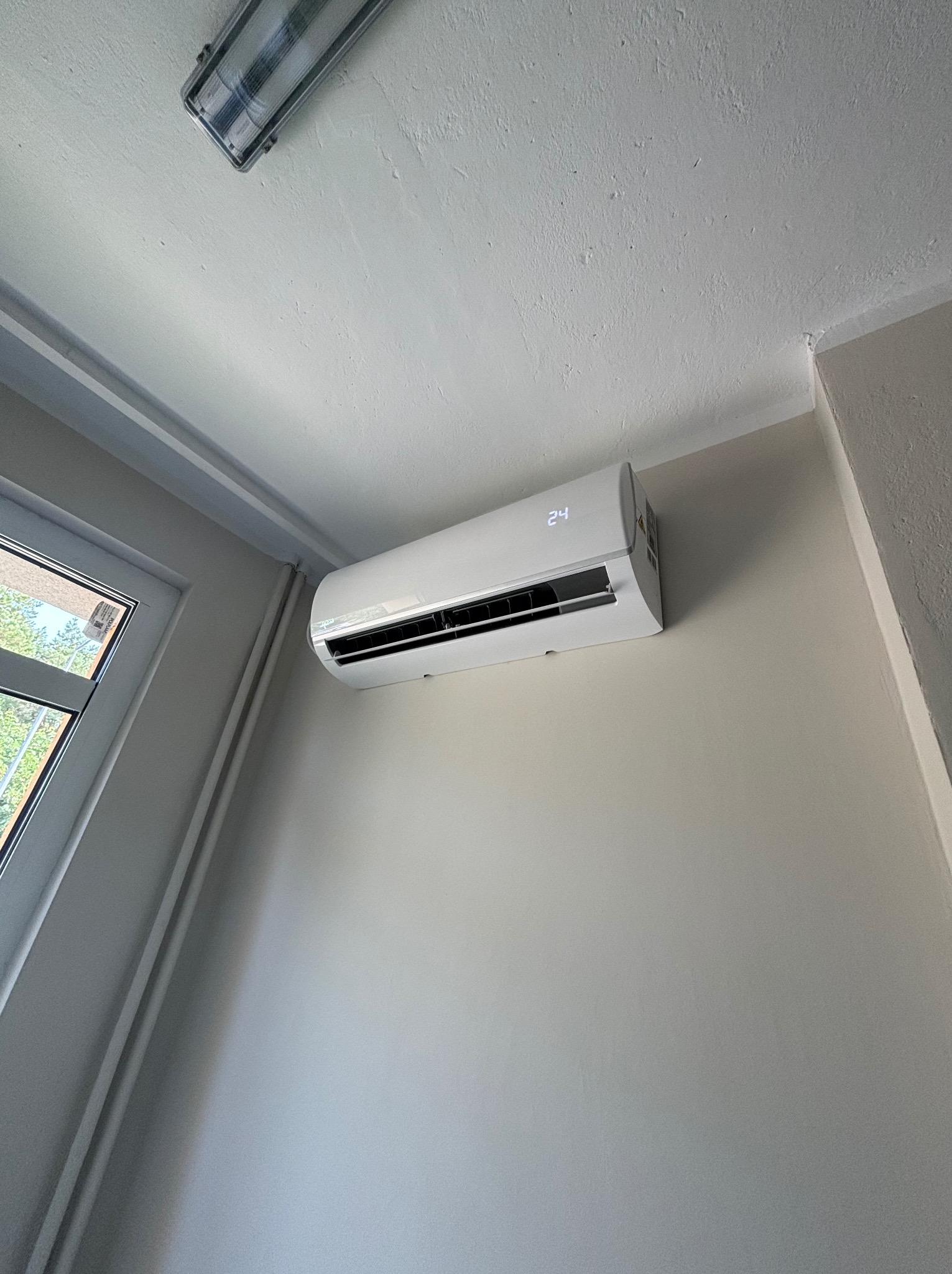 Biała jednostka klimatyzacji na ścianie w pomieszczeniu, widoczny wyświetlacz z temperaturą 24 stopnie, obok okno z widokiem na zieleń.