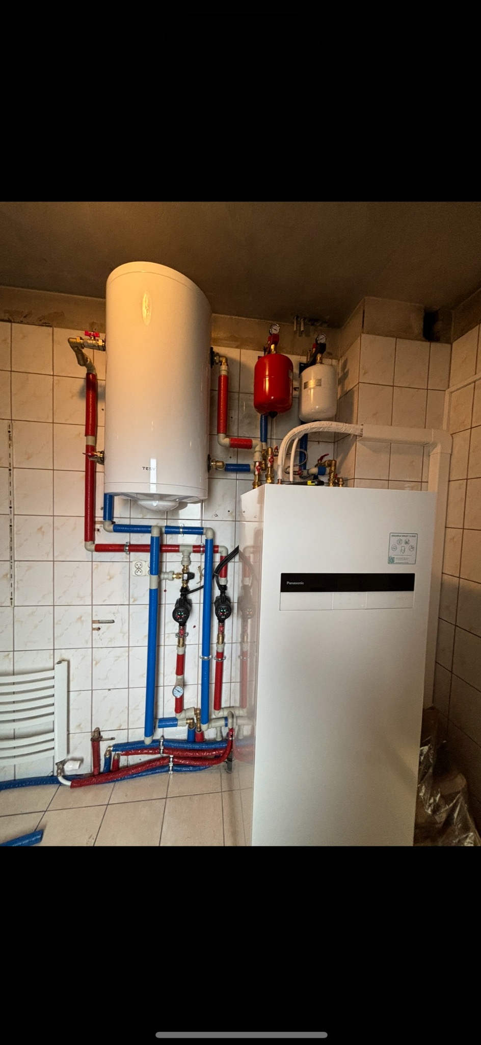 Biały bojler z podłączonymi rurami w kolorach czerwonym i niebieskim, obok biała jednostka pompy ciepła, całość zamontowana w pomieszczeniu wyłożonym płytkami.