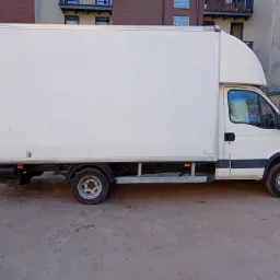 Biały samochód dostawczy z zabudową kontenerową zaparkowany na placu, widok z boku, w tle budynki mieszkalne.